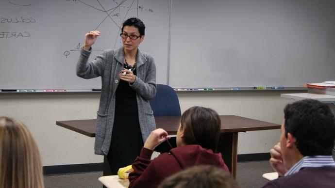 Maria Pia Paganelli stands at front of classroom, holding dry erase marker, teaching students in front of a whiteboard