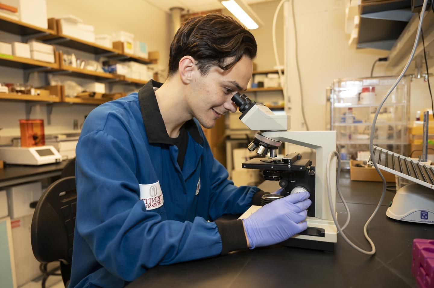 A student in lab coat and gloves looks through a microscope