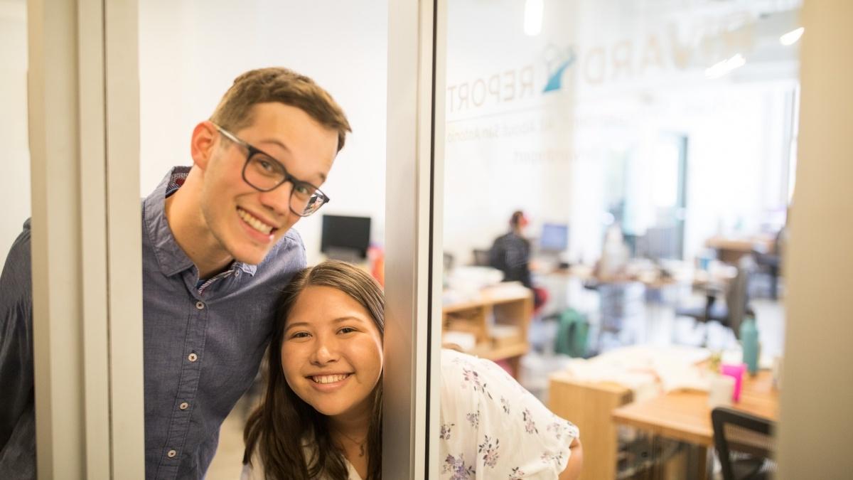 Two students peaking through glass door at Rivard Report